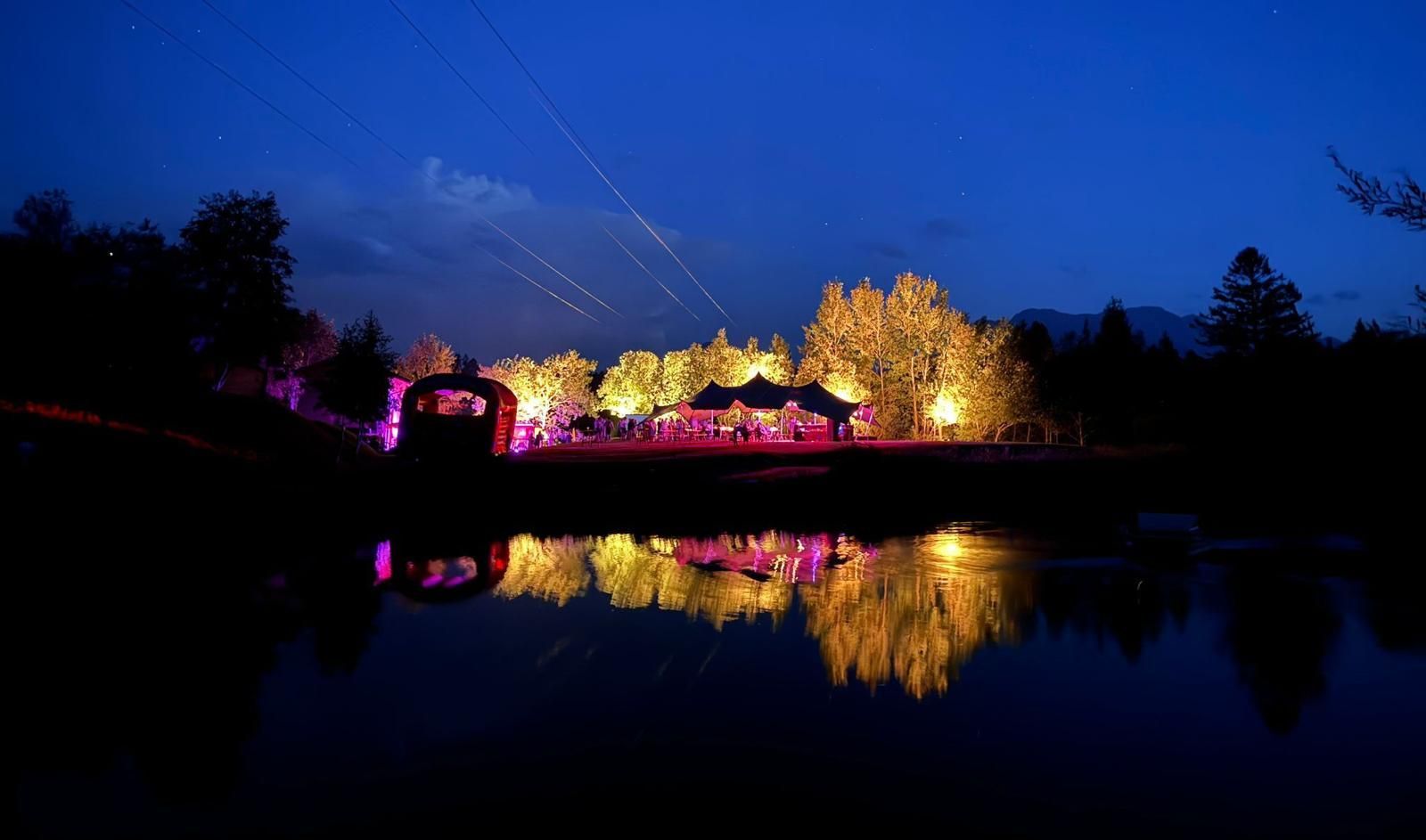 Eine Brücke über einen See bei Nacht mit Bäumen und Lichtern, die sich im Wasser spiegeln.