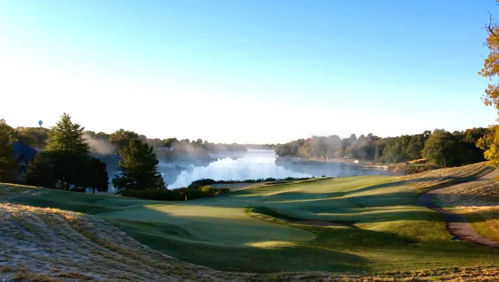 Golf course green overlooking a misty lake, surrounded by trees under a clear sky.