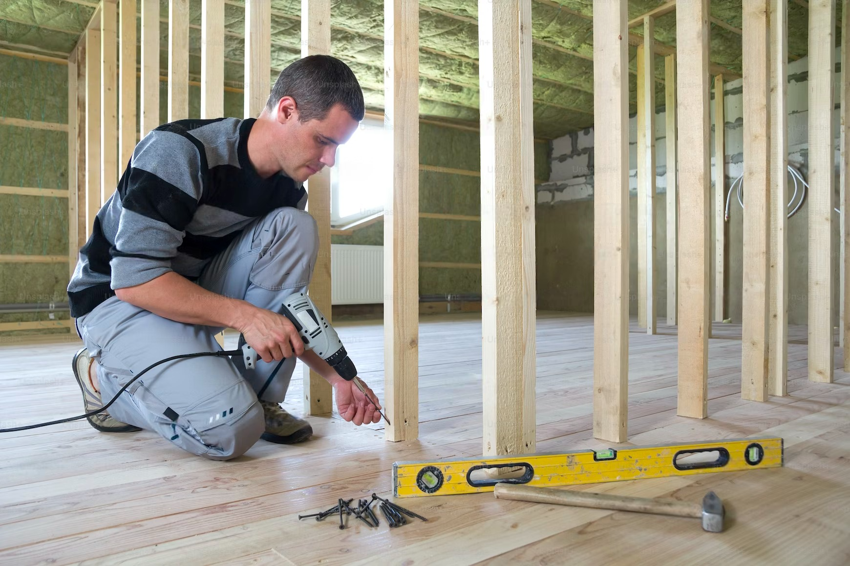 Man using a drill on a wooden frame, holding a level and a hammer, indoors.
