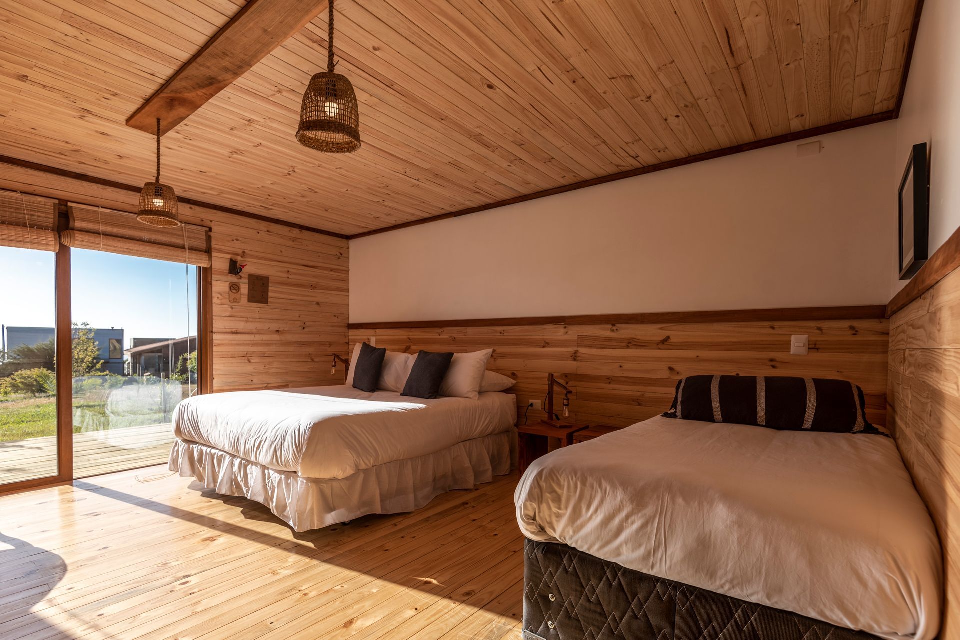 Dormitorio con paneles de madera, con dos camas, grandes puertas de vidrio y una luz decorativa.