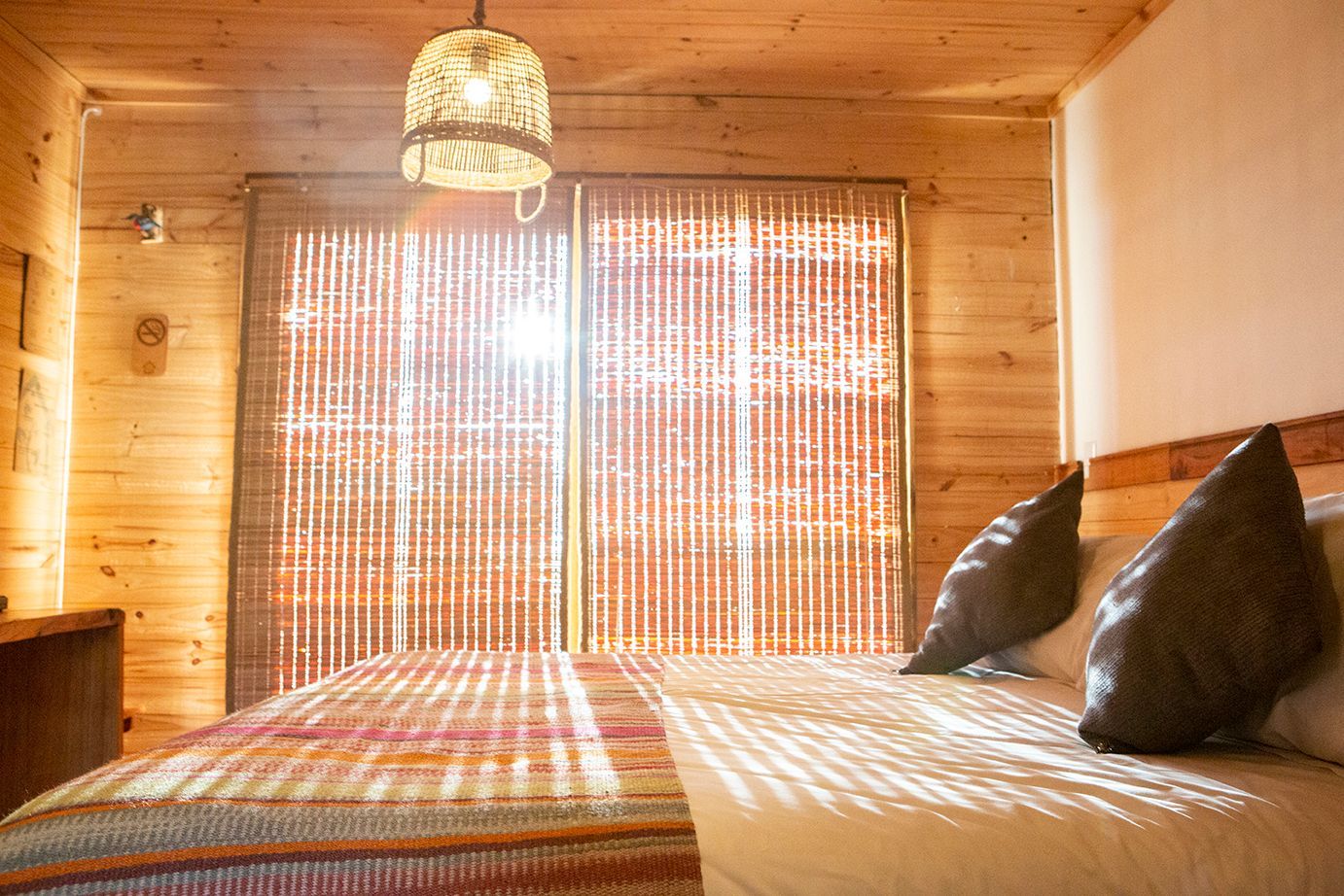 Dormitorio con paredes de madera, cama, cortinas tejidas y lámpara colgante.
