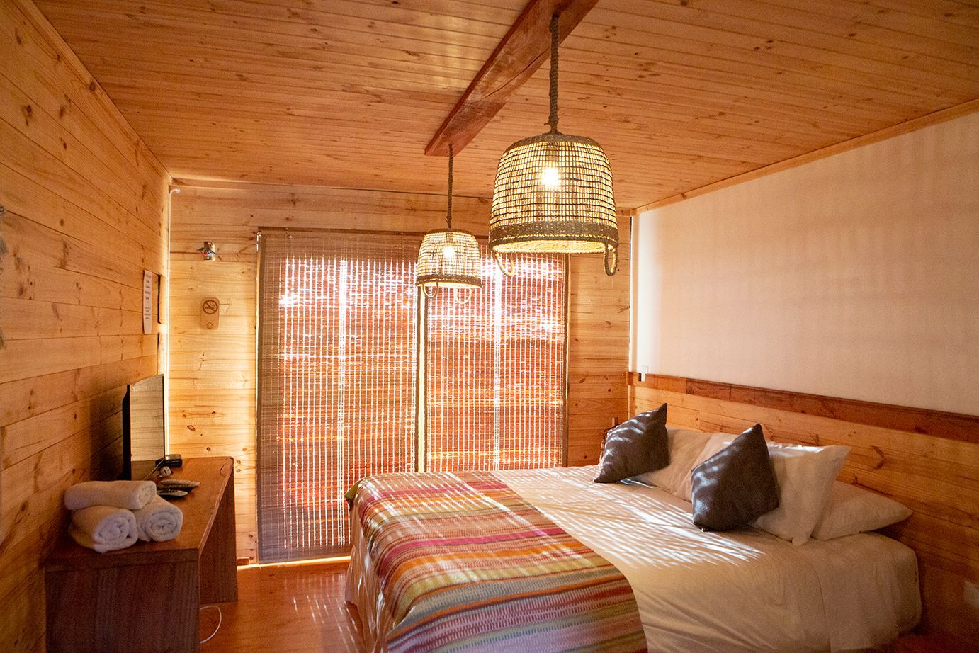 Dormitorio de madera con cama, toallas, ventana y lámparas colgantes.