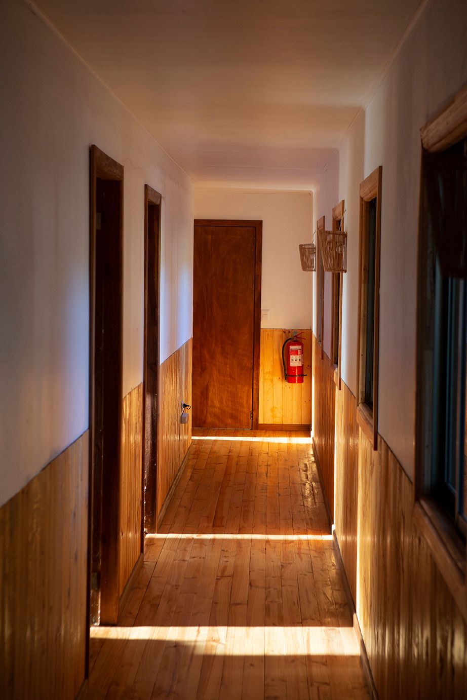 Pasillo estrecho con paneles y puertas de madera; la luz del sol entra a raudales. Hay un extintor de incendios montado.