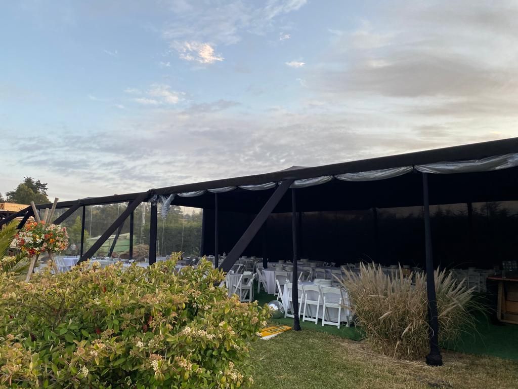 Carpa para eventos al aire libre con paredes de tela negra, sillas blancas y mesas sobre un césped.