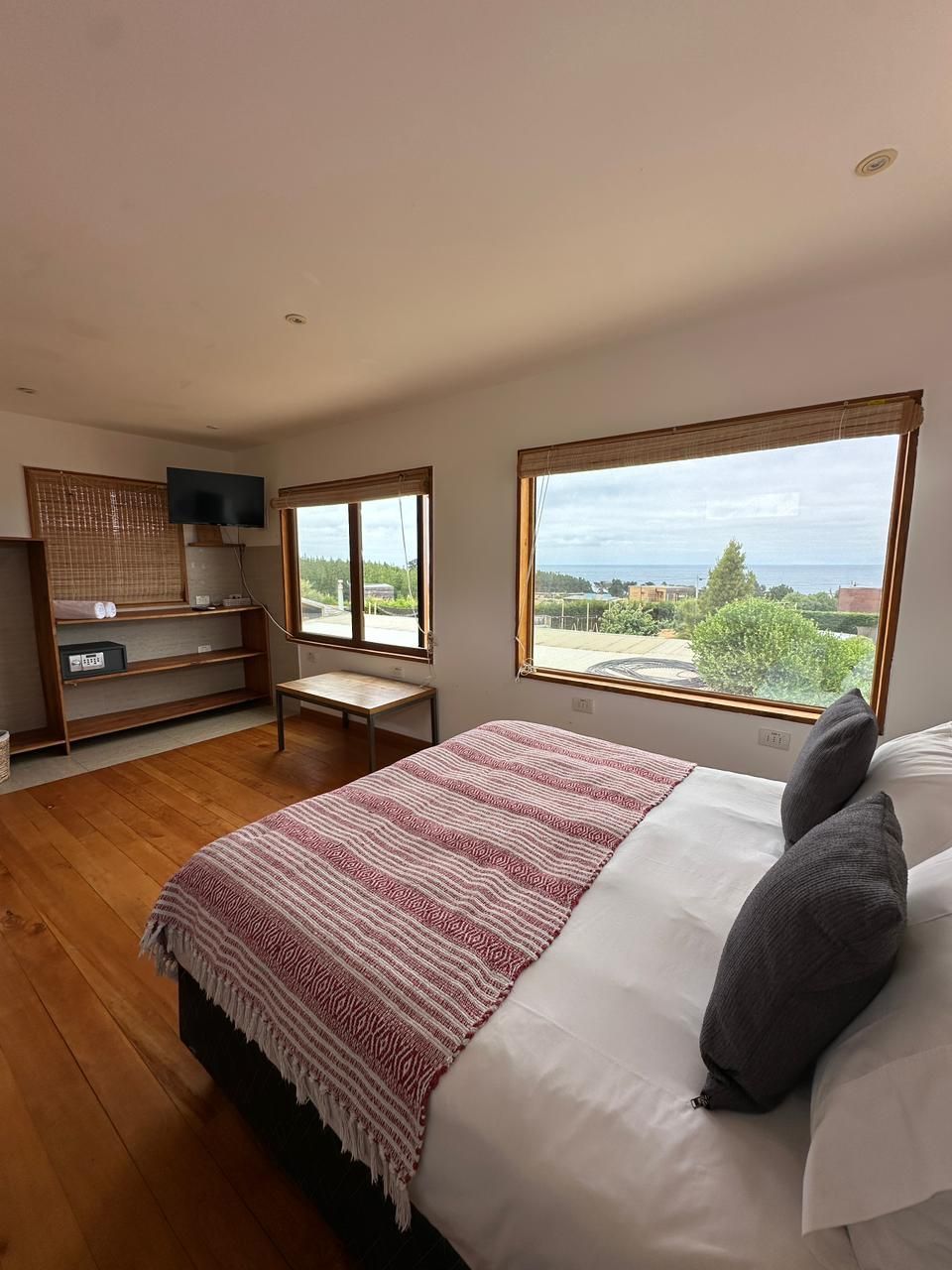 Dormitorio con cama, amplios ventanales y vistas. Suelo de madera, manta roja y blanca y un pequeño televisor.