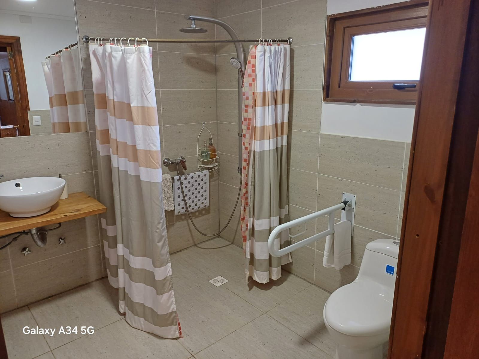 Baño accesible con ducha, inodoro y lavabo. Paredes de azulejos beige, barra de apoyo y cortinas de ducha.