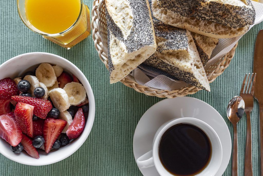 Desayuno: bol de frutas, pan de molde, zumo de naranja, café y cubiertos sobre un mantel individual color verde azulado.