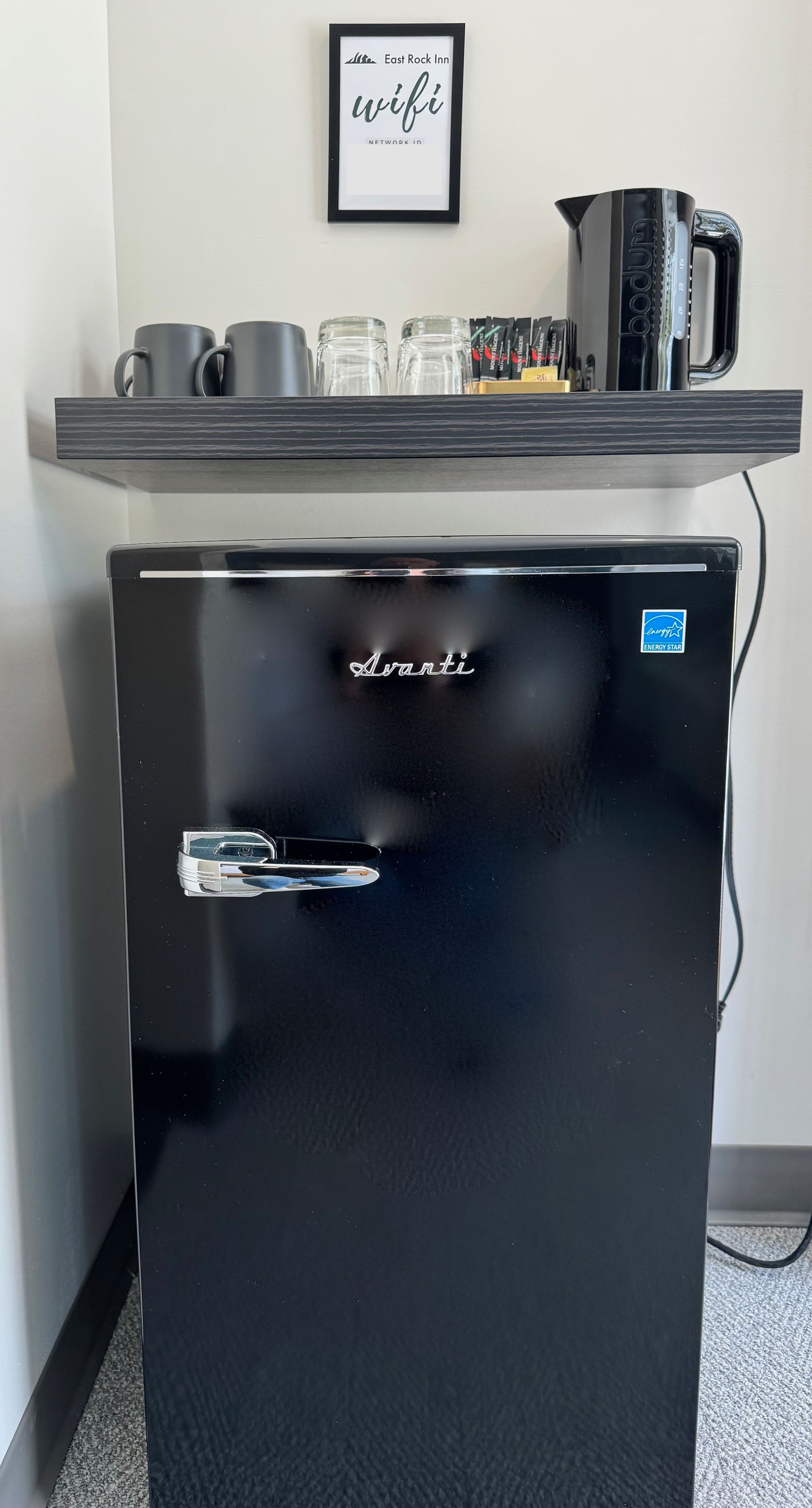 A black refrigerator is sitting in a corner of a room next to a shelf.