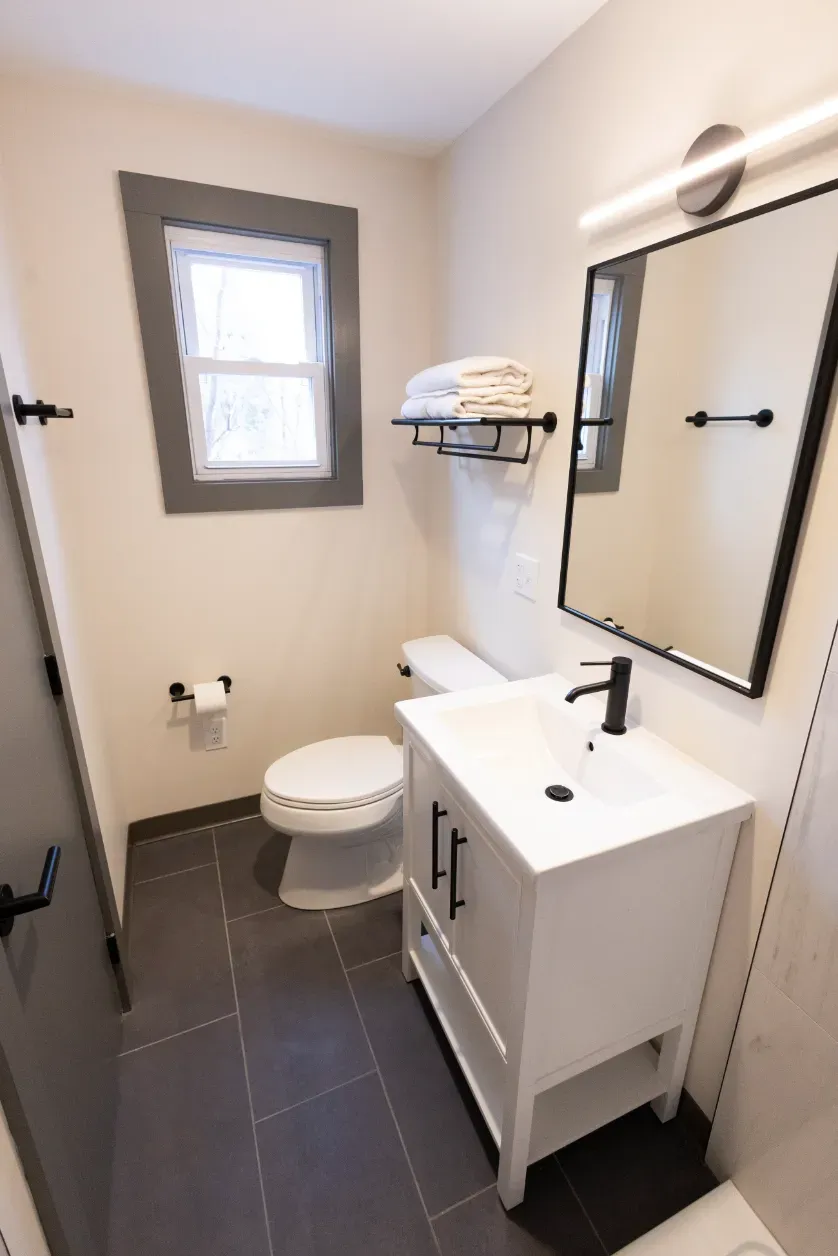 A bathroom with a toilet , sink , mirror and window.