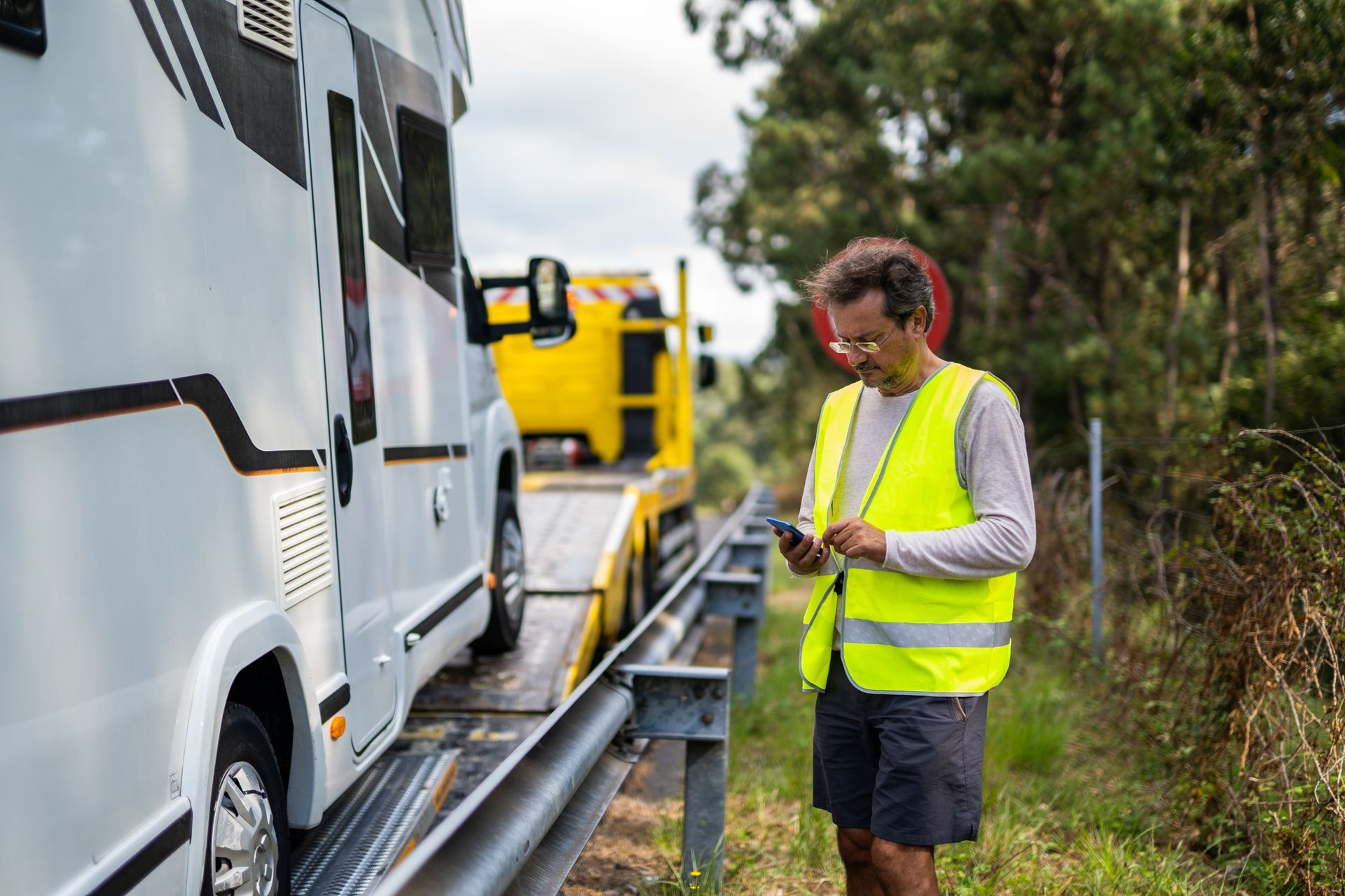 Man in safety vest calls for help next to a broken-down RV being towed on the roadside.