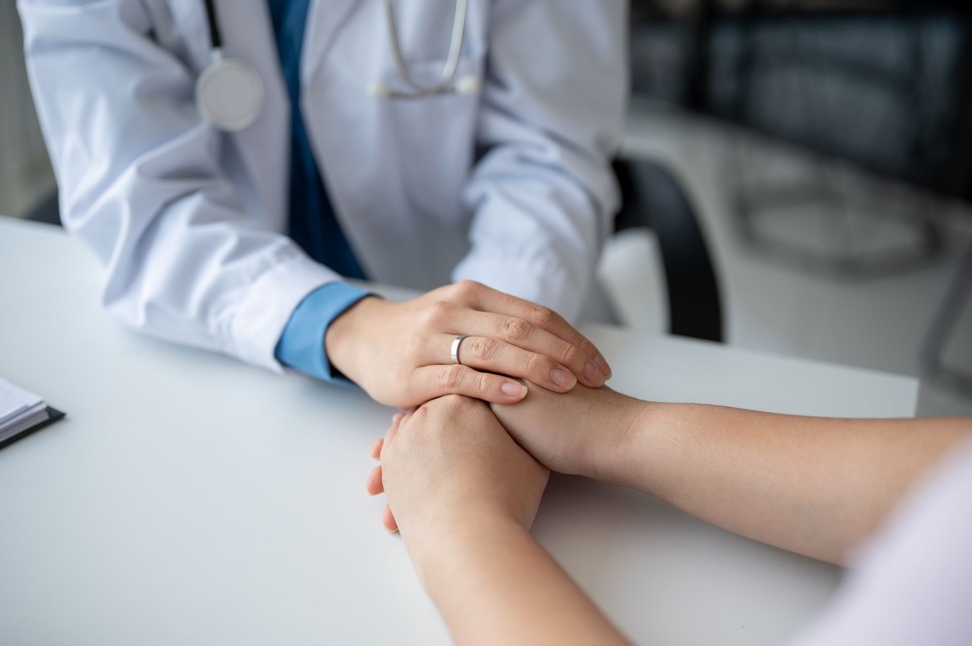 Doctor's hands gently holding a patient's hands over a white desk. A stethoscope hangs around the doctor’s neck.