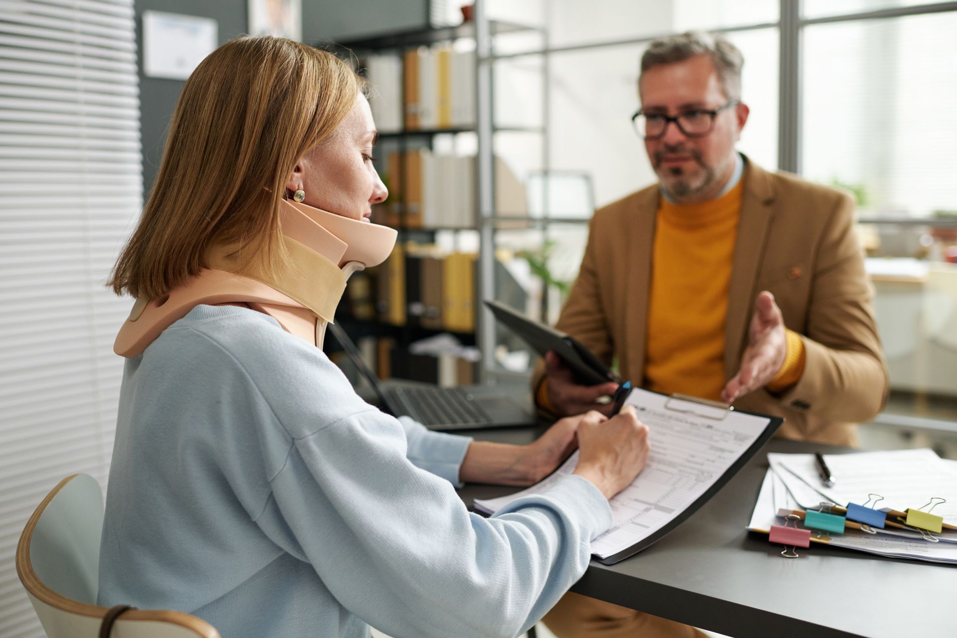 Woman wearing neck brace signs documents with a man in an office.