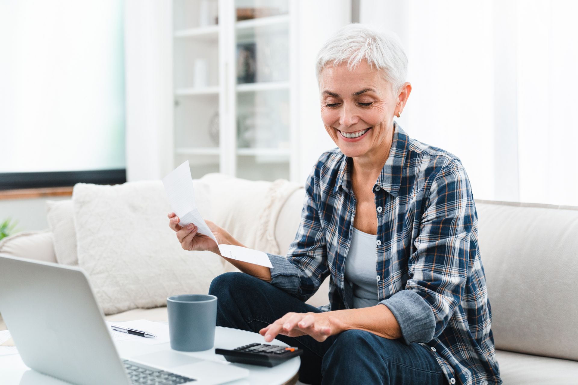 Woman calculating bills on a laptop, smiling. Sitting on a couch with a coffee cup.