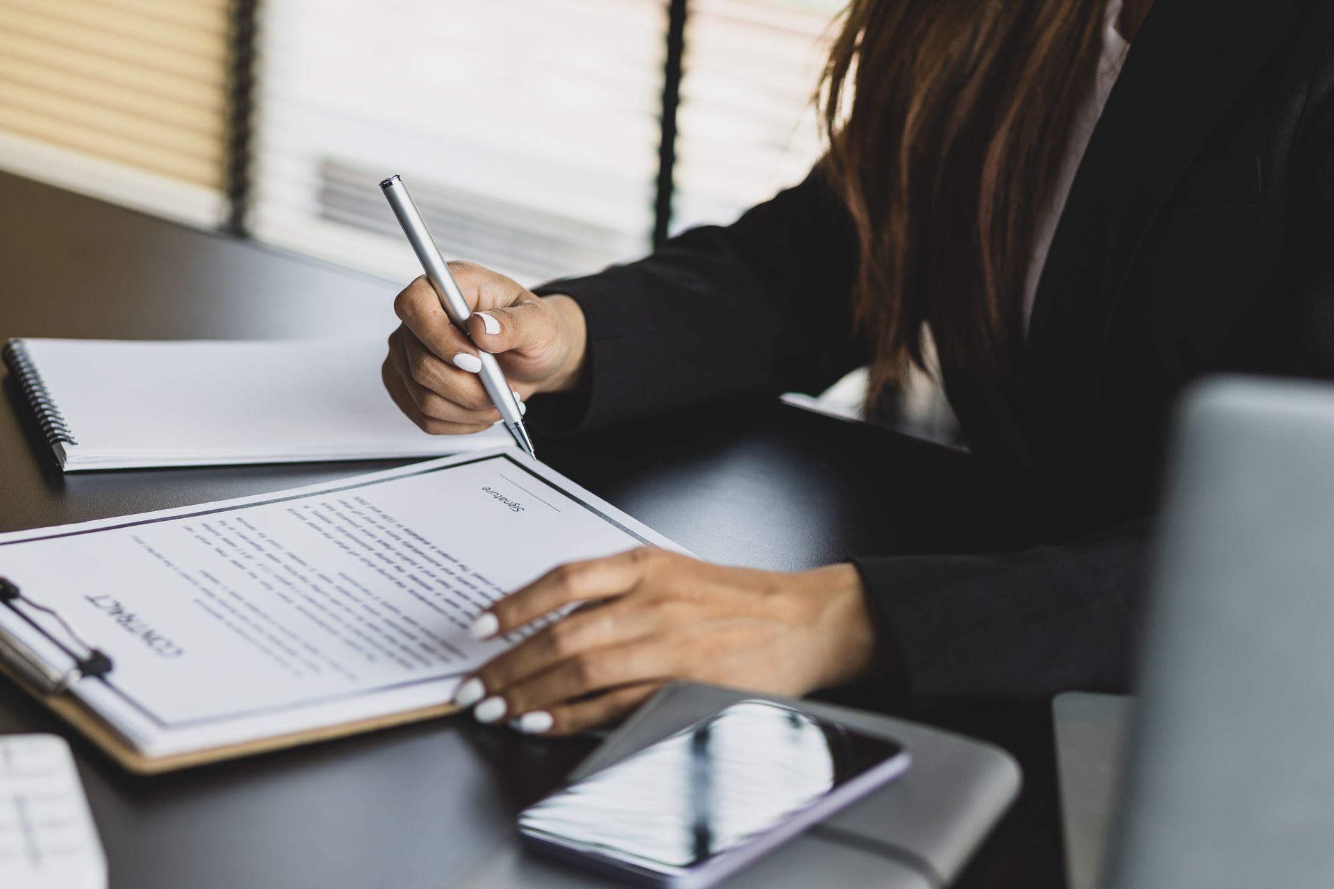 Woman in black blazer signing a contract at a desk with a phone and laptop.