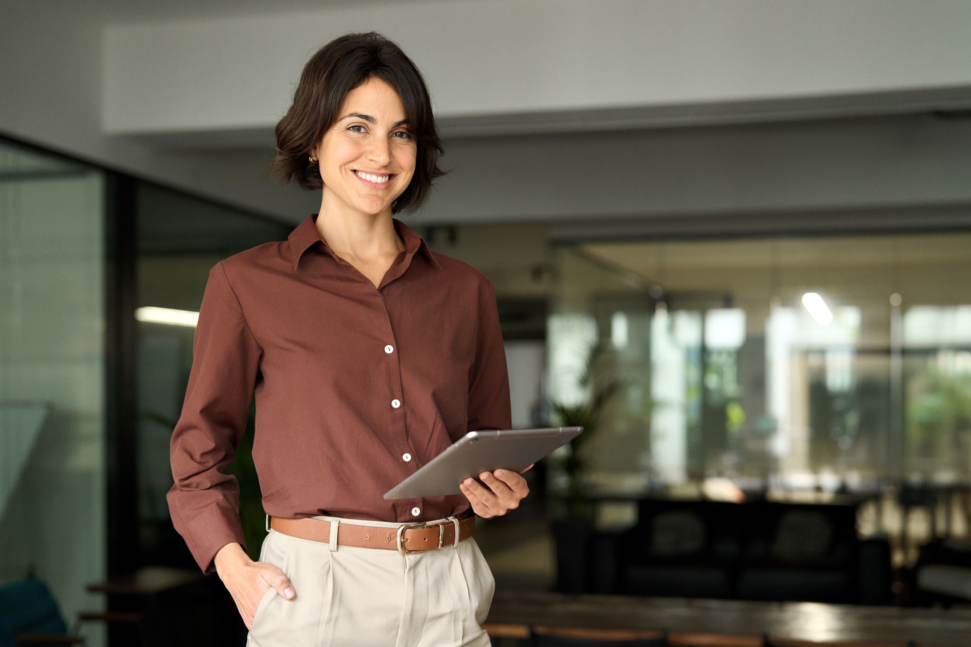 Woman in brown shirt and tan pants holds tablet, smiles in office setting.