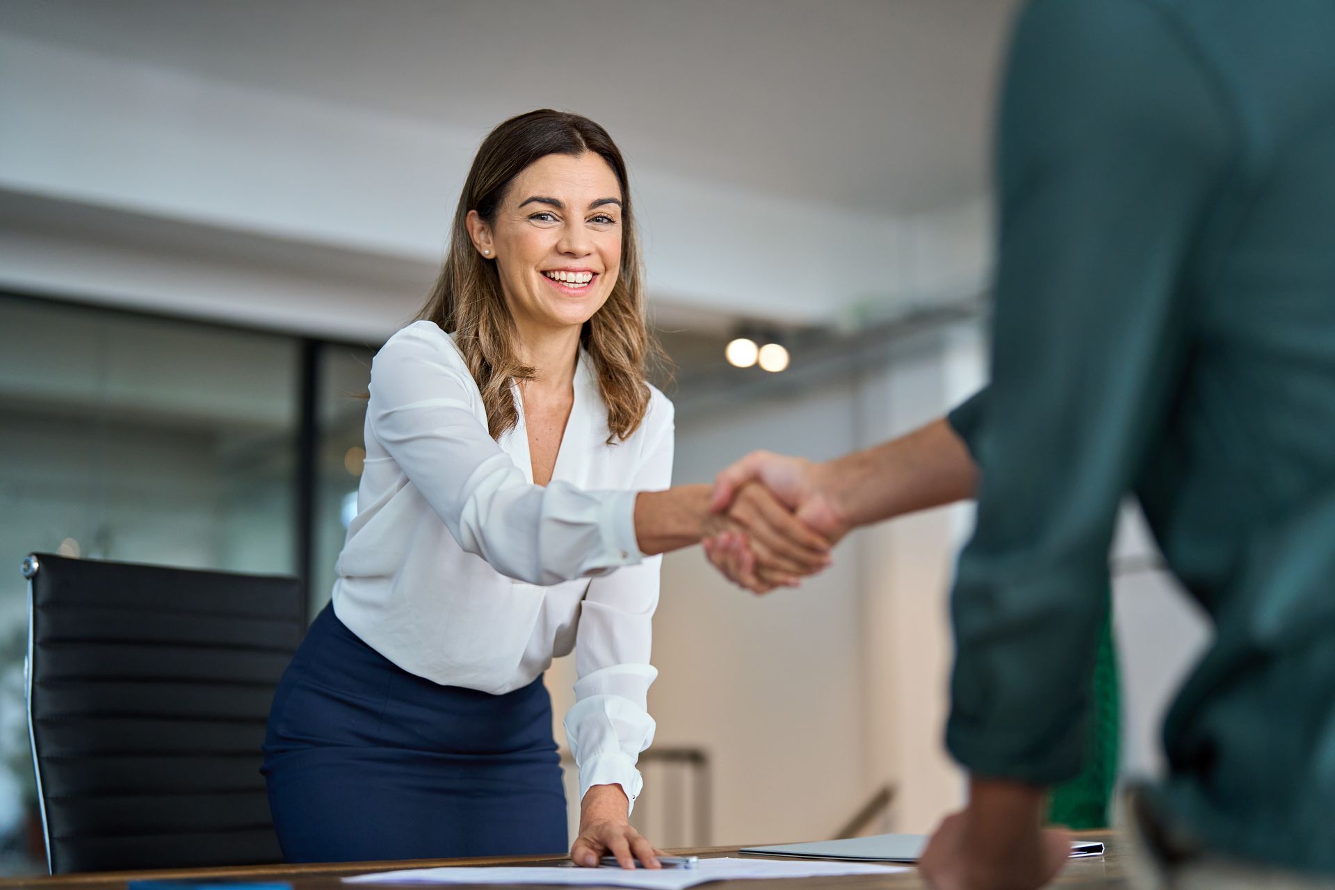 Woman in a white blouse and blue skirt shaking hands with another person at a desk in an office.