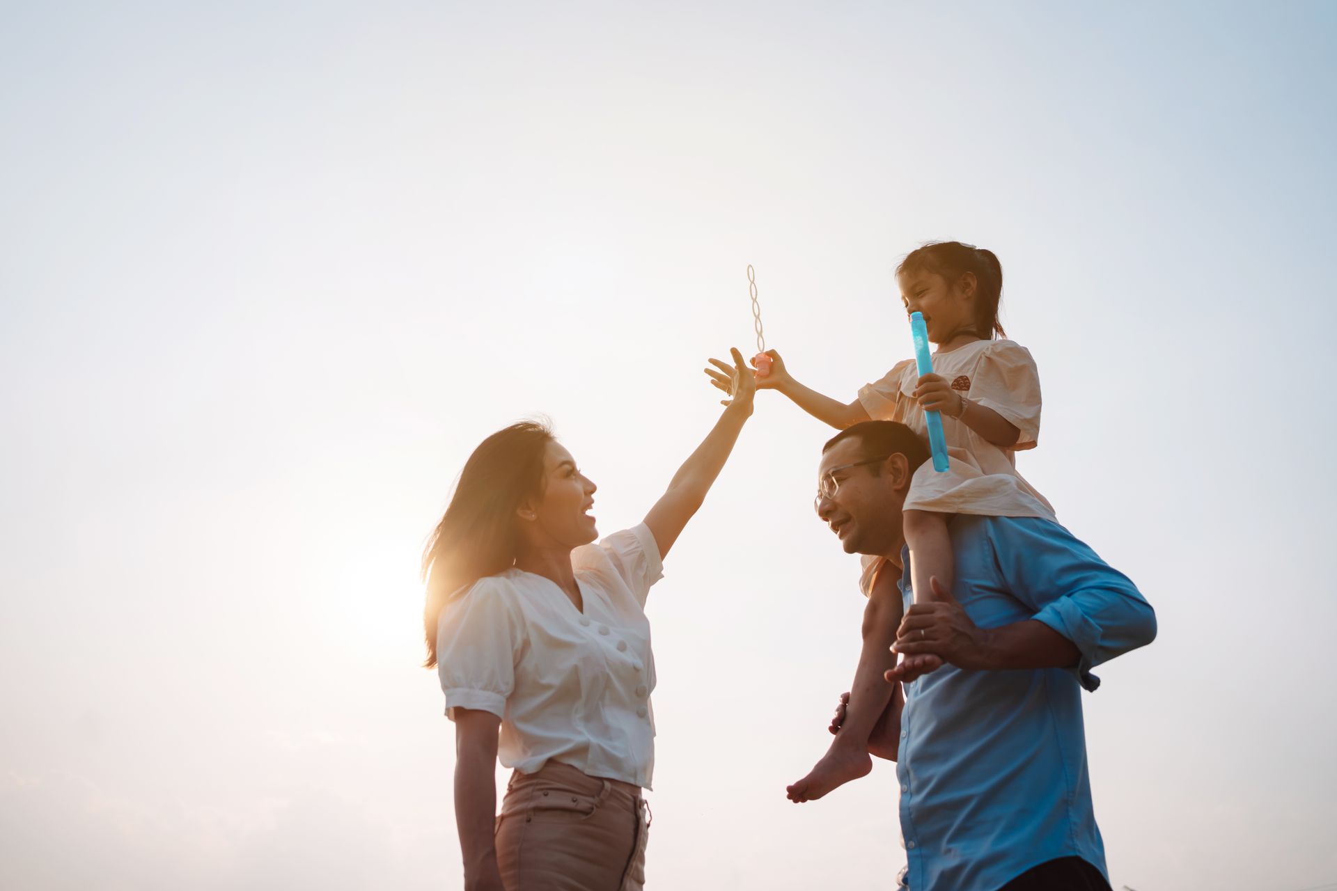 Family at sunset: father carries child on shoulders, mother high-fives the child with bubbles.