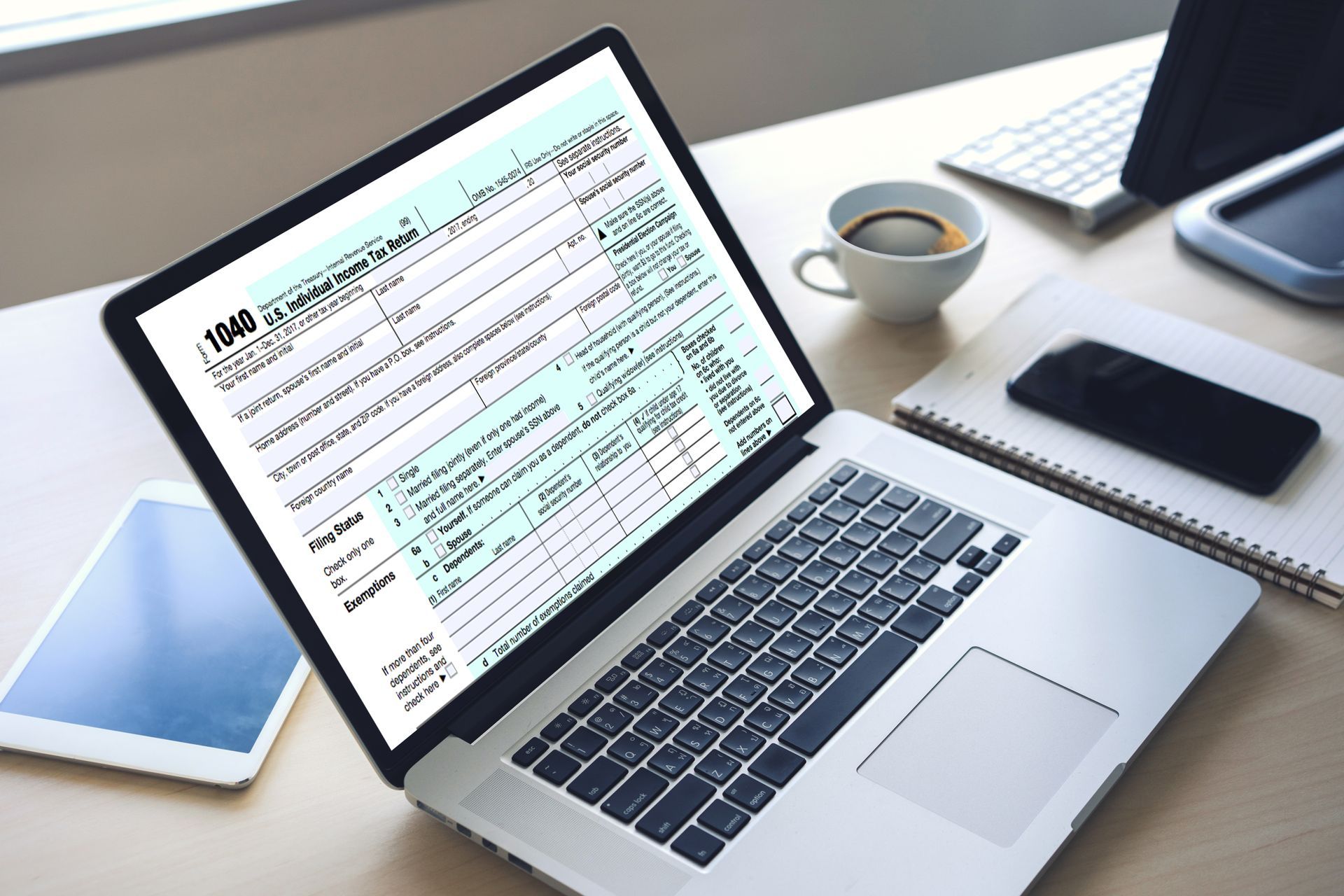 Laptop displaying a 1040 tax form; coffee cup, notebook, tablet, and smartphone on a desk.