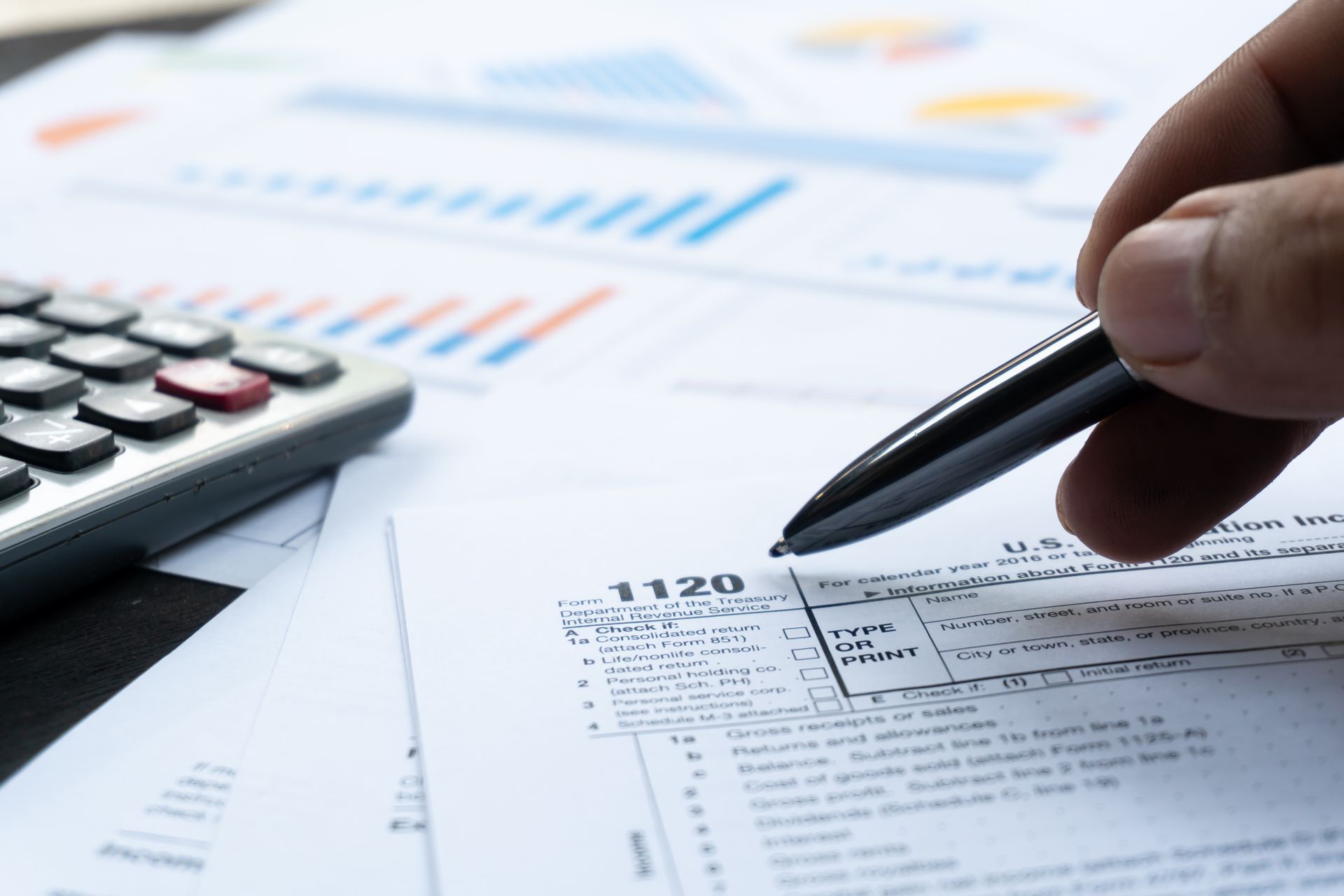 Hand holding pen over 1120 tax form, calculator, and financial reports.