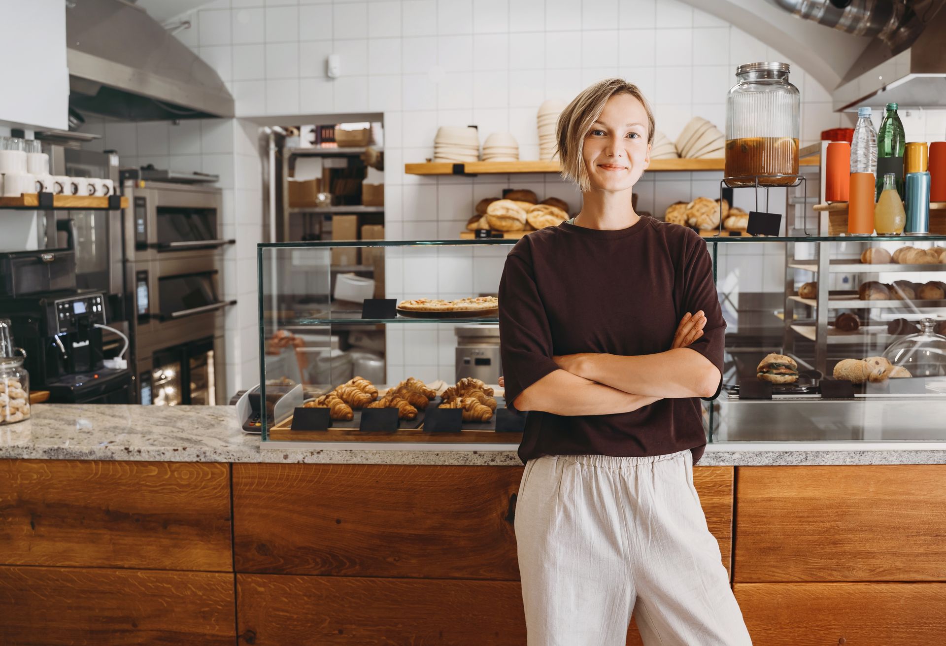 Woman with crossed arms in a bakery; behind her are baked goods in a display case and on shelves.