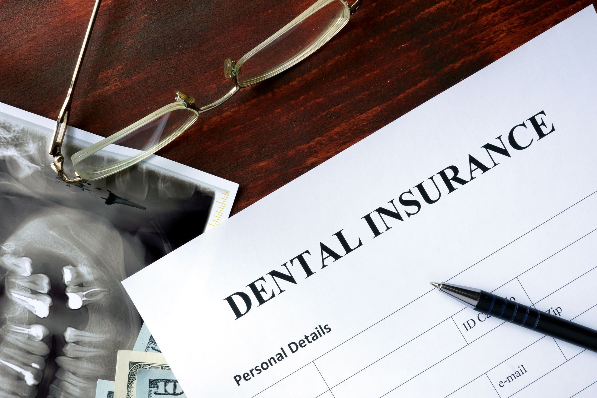 Dental insurance form, glasses, pen, dental X-ray, and money on a wooden surface.