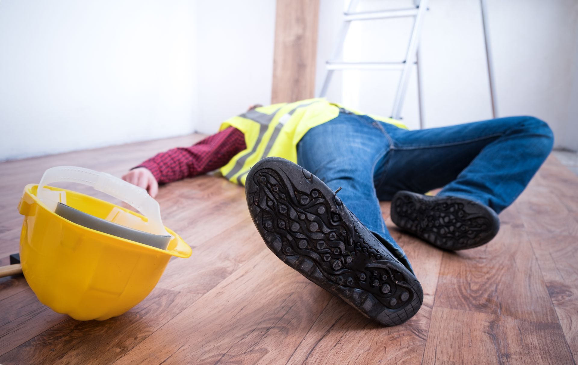 Construction worker lying on floor, wearing a yellow safety vest, hard hat nearby, ladder in background.