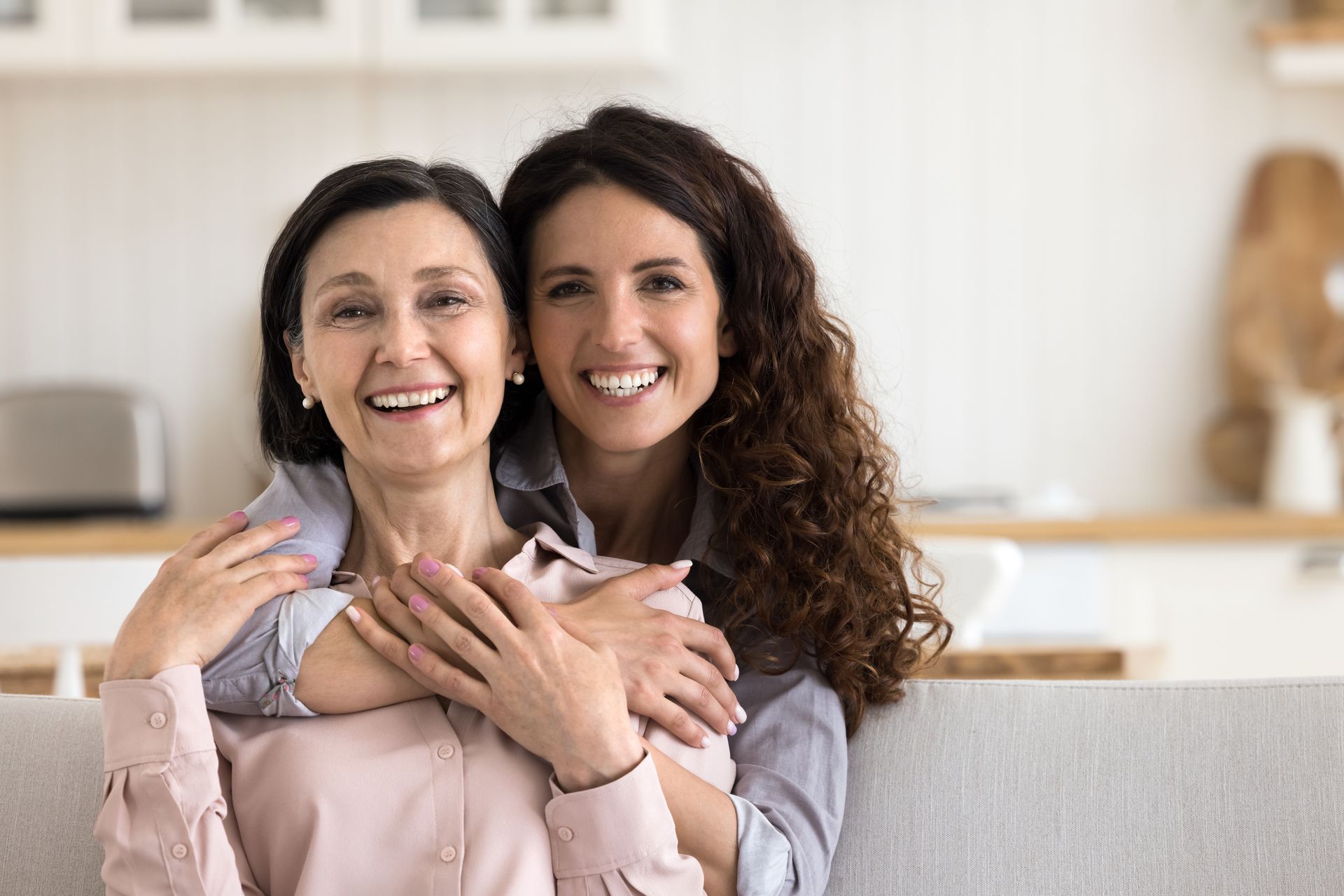 Woman embraces another smiling woman from behind, indoors.