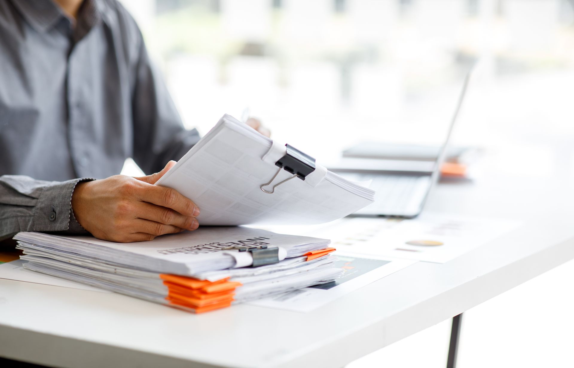Person in grey shirt sorts through a stack of documents clipped together, working at a desk with a laptop.