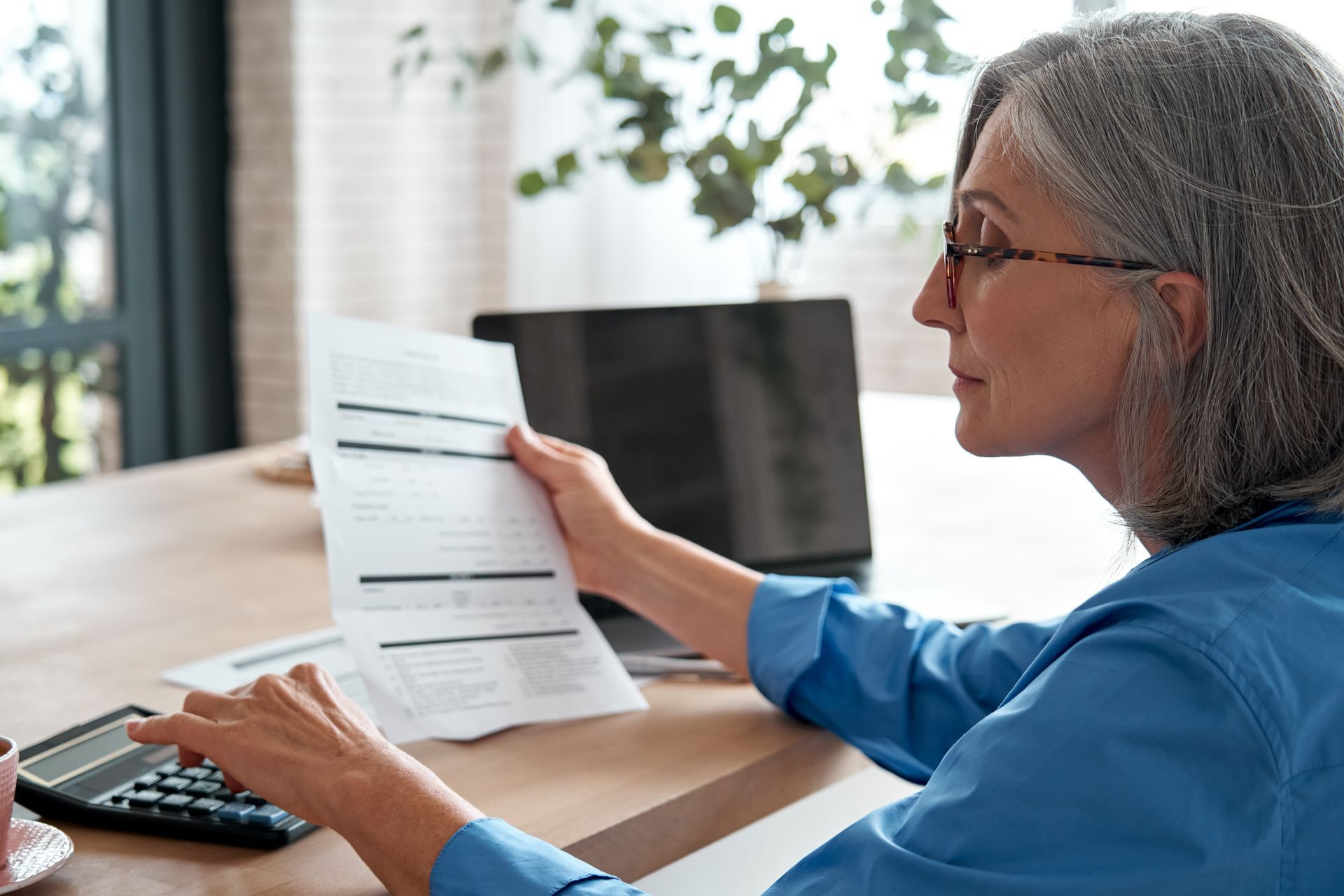 Woman with glasses looks at papers, using a calculator, near a laptop and coffee cup.