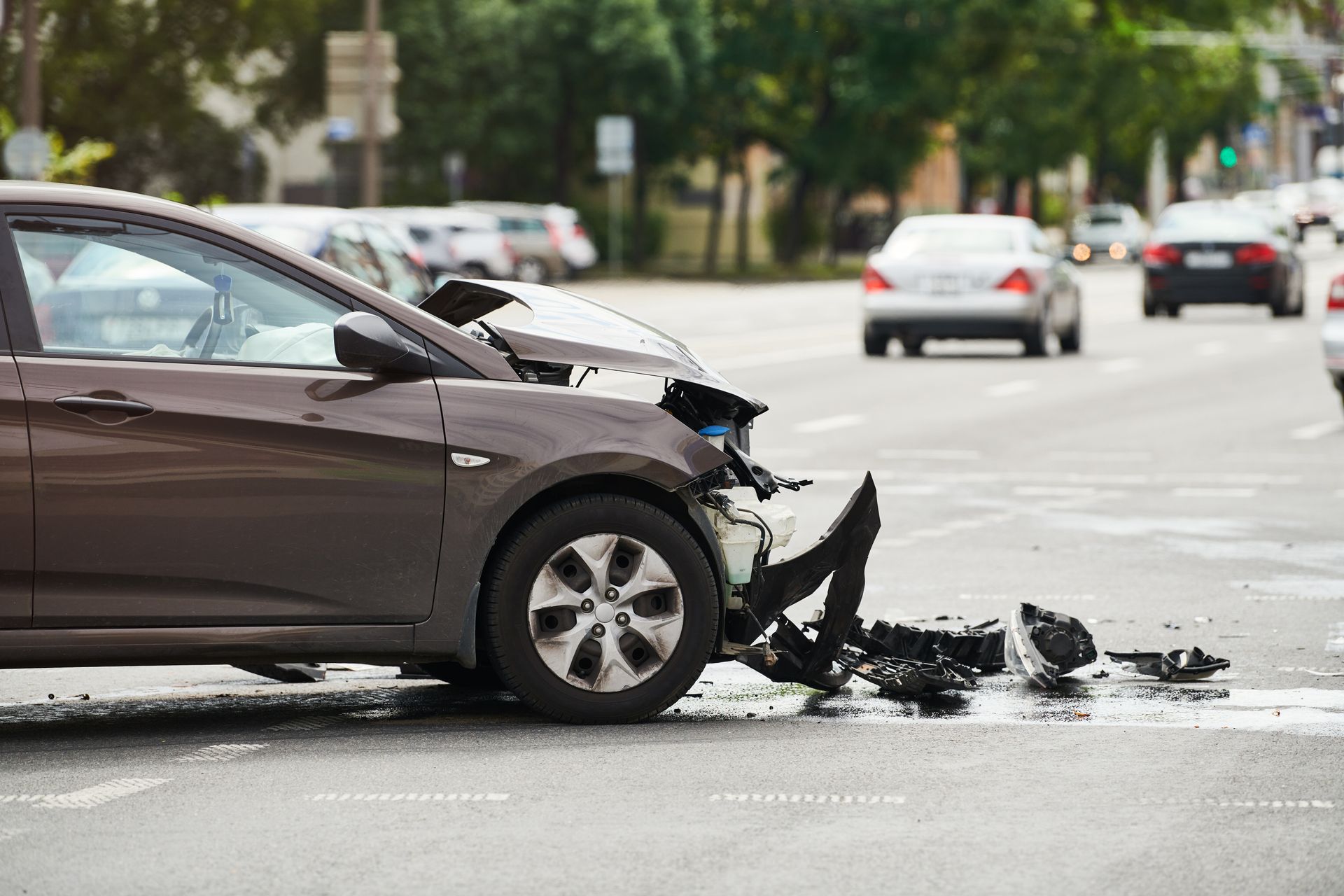 Brown car damaged in a traffic accident at an intersection; debris scattered on the road, other cars in the background.