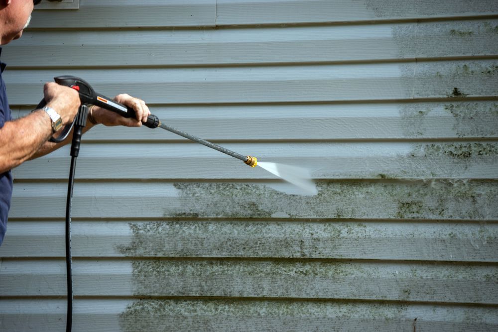Person using a pressure washer to clean mold off of a light-colored house siding.