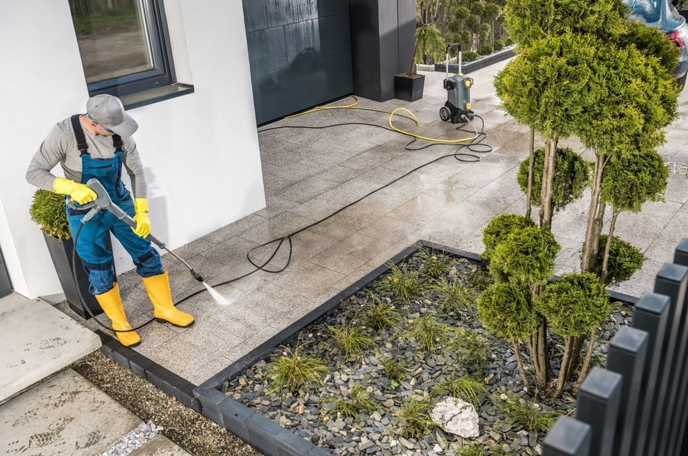 Person power washing a concrete patio next to a garden bed and a home.