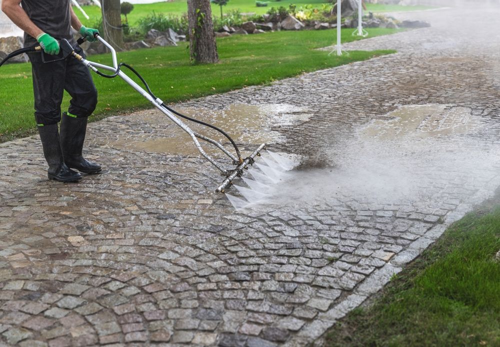 Person power washing a cobblestone walkway with a silver pressure washer. Green grass and trees are in the background.