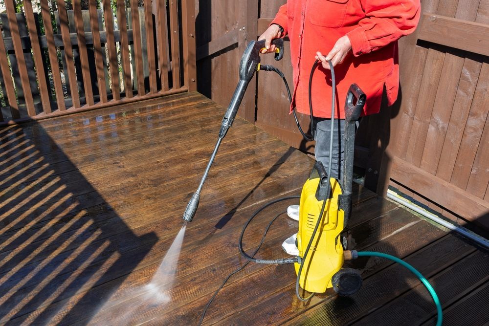 Person pressure washing a wooden deck with a yellow machine and silver wand.