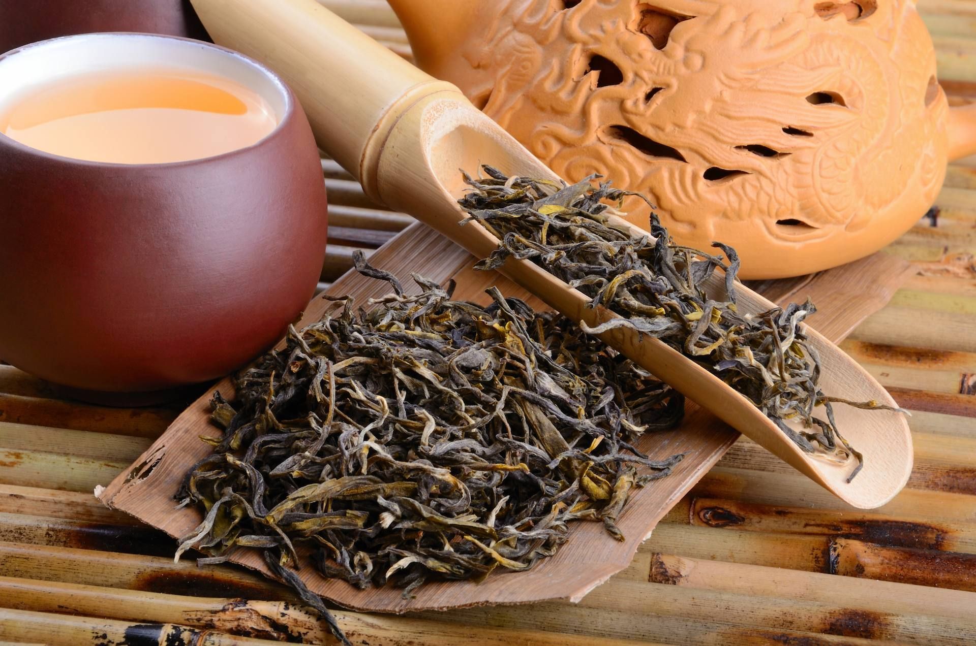 Tea leaves on a bamboo scoop, with a cup and teapot, all on a bamboo mat.