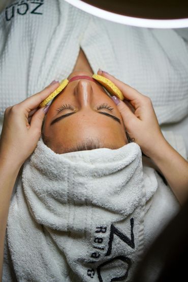 Woman receiving facial treatment, relaxed and serene. Beige sponges on eyes, white robe, towel.