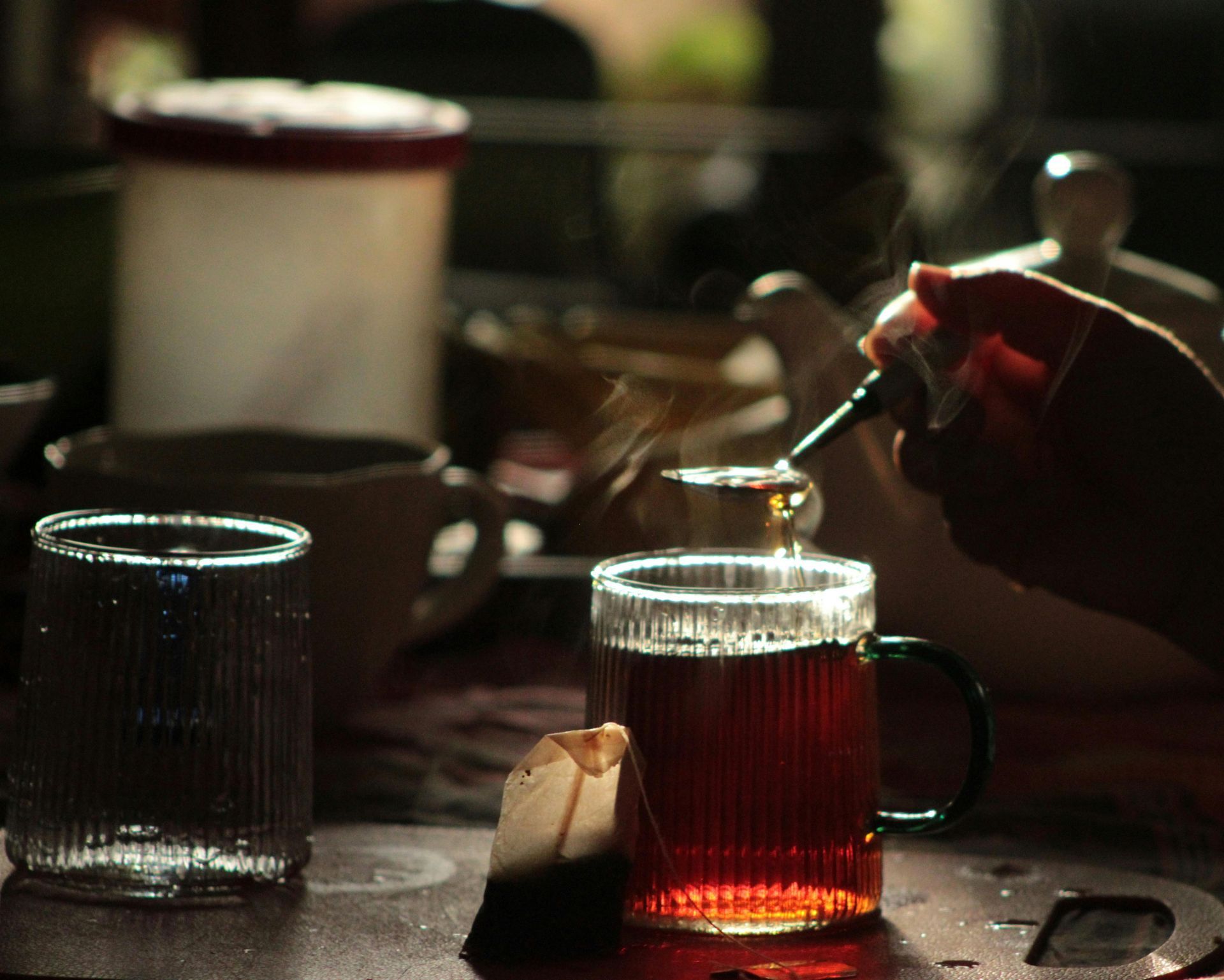 A hand pouring tea into a glass mug with a tea bag; other items on a tabletop.