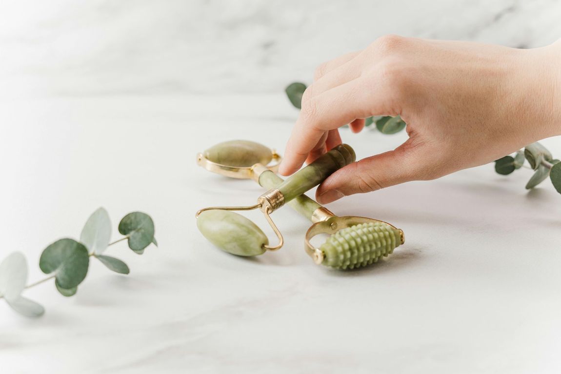 Hand holding a jade facial roller on a white marble surface next to eucalyptus leaves.