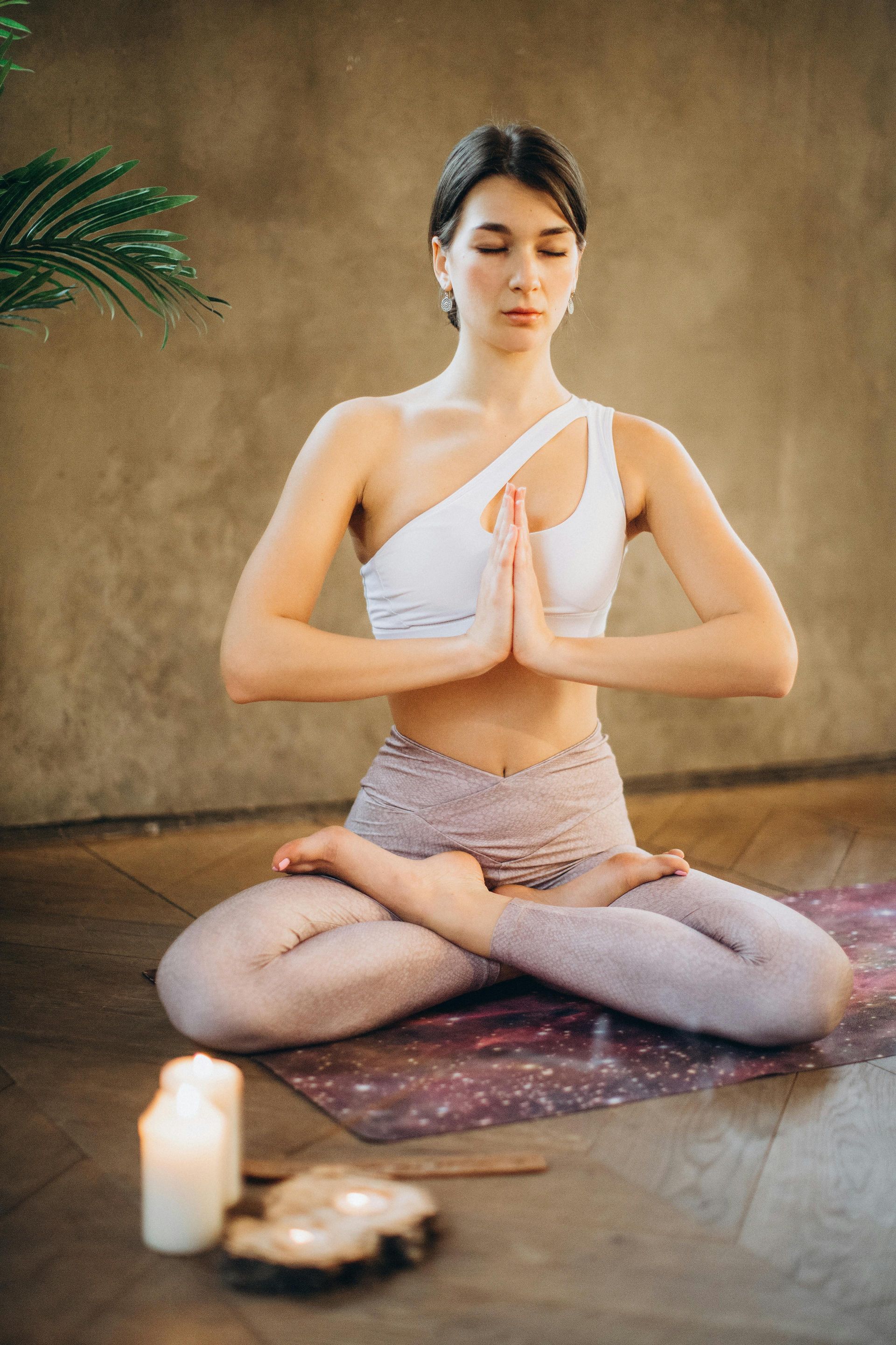 Woman in lotus pose, hands in prayer, eyes closed, wearing white top, purple leggings, meditating indoors.