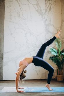Woman in a yoga bridge pose, leg extended upward, in a light-filled room with marble wall, on a blue mat.