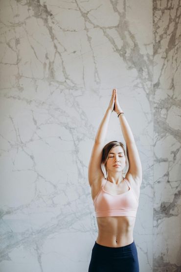 Woman in yoga pose, arms raised overhead, palms touching, eyes closed, pink top, marble wall.