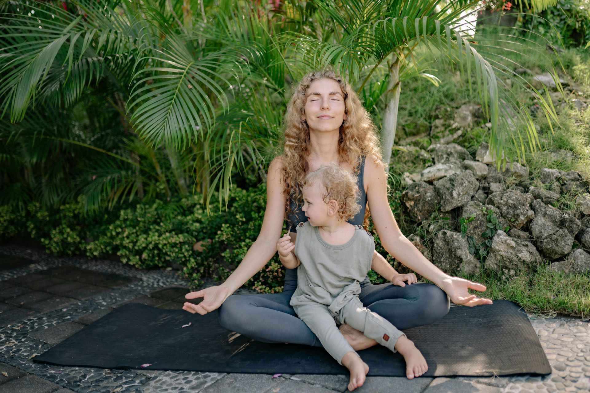 Woman with curly hair meditating outdoors with a small child, palms up.