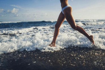 Woman in white bikini runs along the foamy shore of a blue ocean.