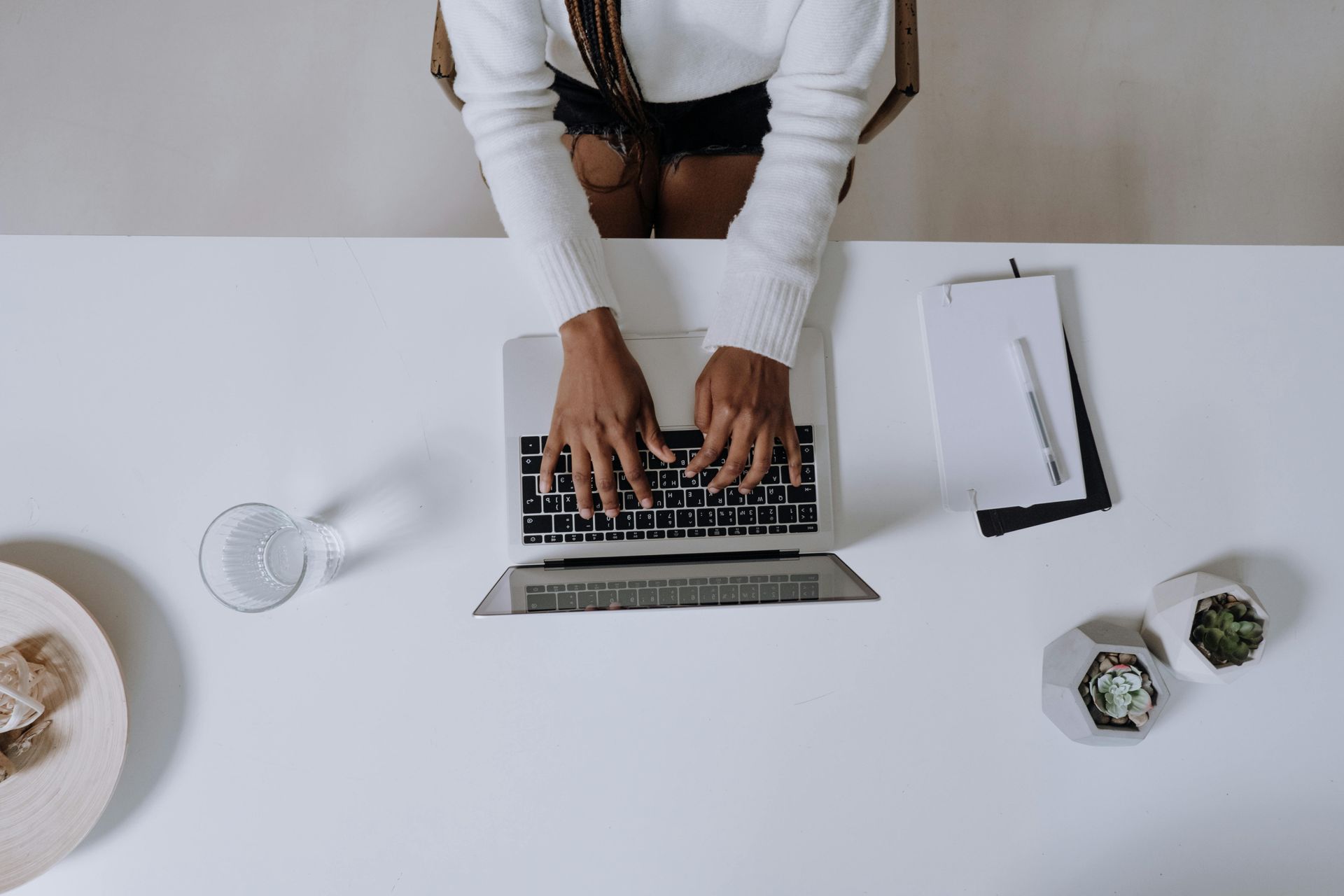 Person typing on a laptop at a white desk; a glass, notepad, and succulent plants are nearby.
