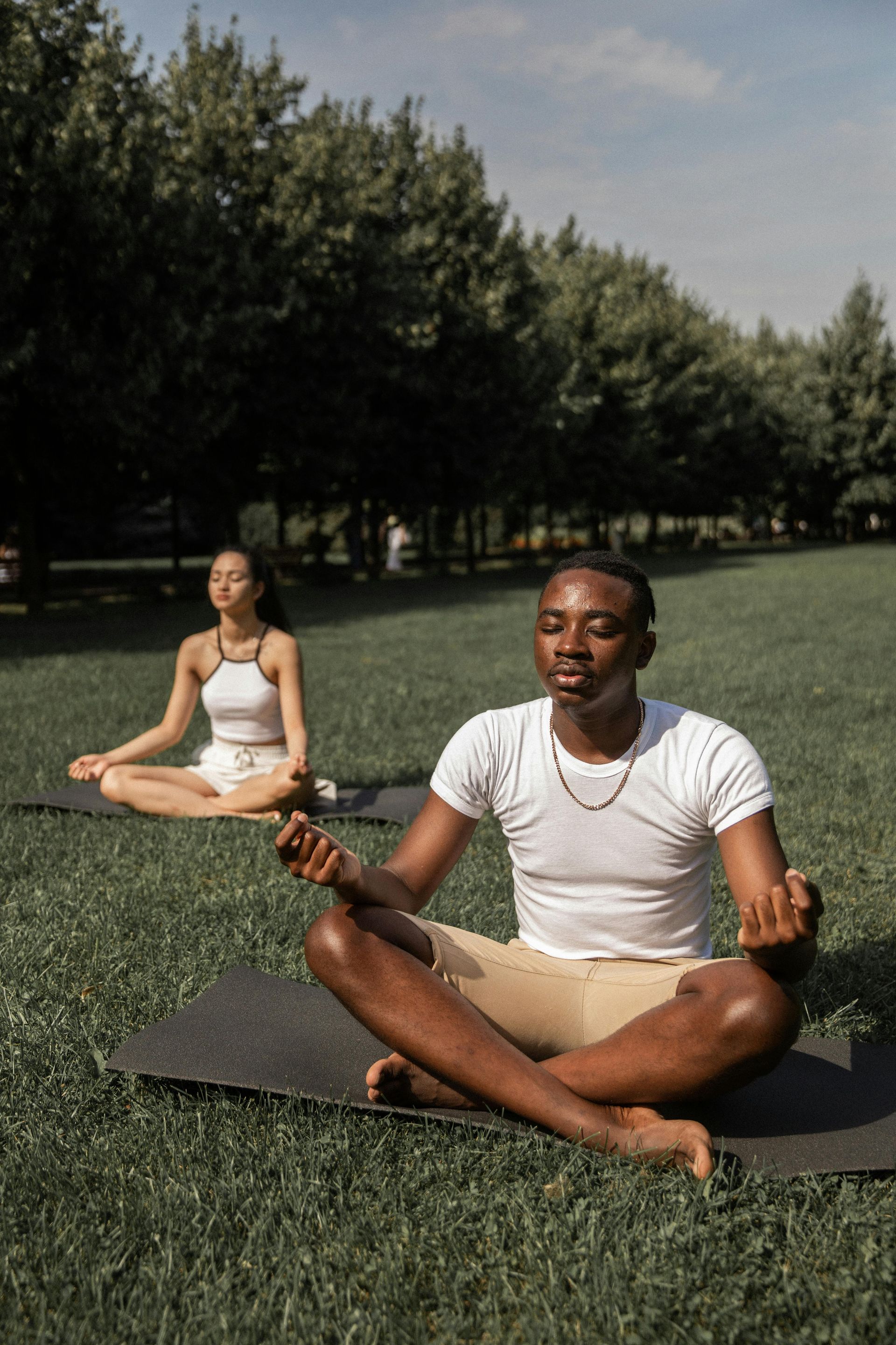Two people meditating on yoga mats in a park. Man in white t-shirt, woman in tank top, both with closed eyes.