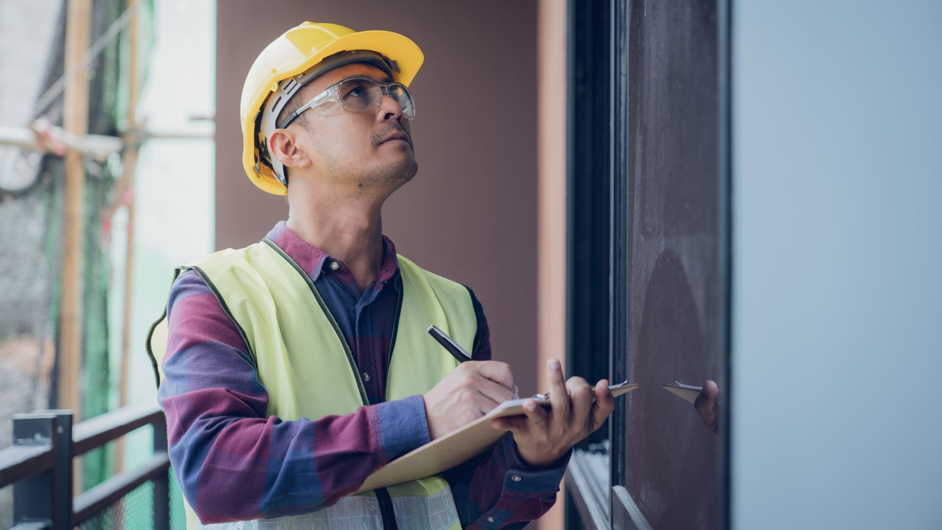 Un ouvrier du bâtiment, portant un casque et un gilet jaunes, inspecte l'extérieur d'un bâtiment et prend des notes.