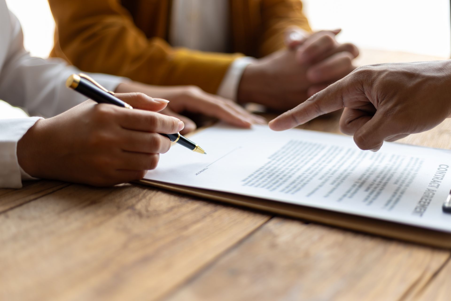 Une personne signe un document, avec une autre personne et une main pointant vers lui sur une table en bois.