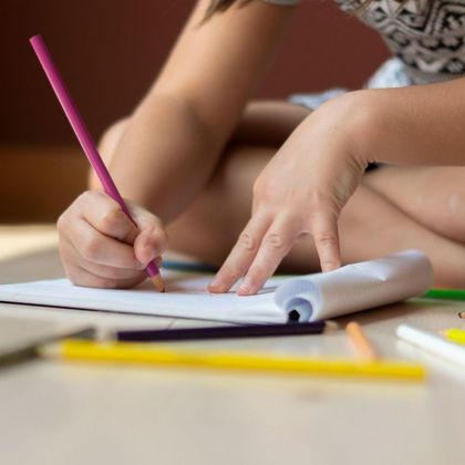 A child is writing on a piece of paper with a purple pencil.