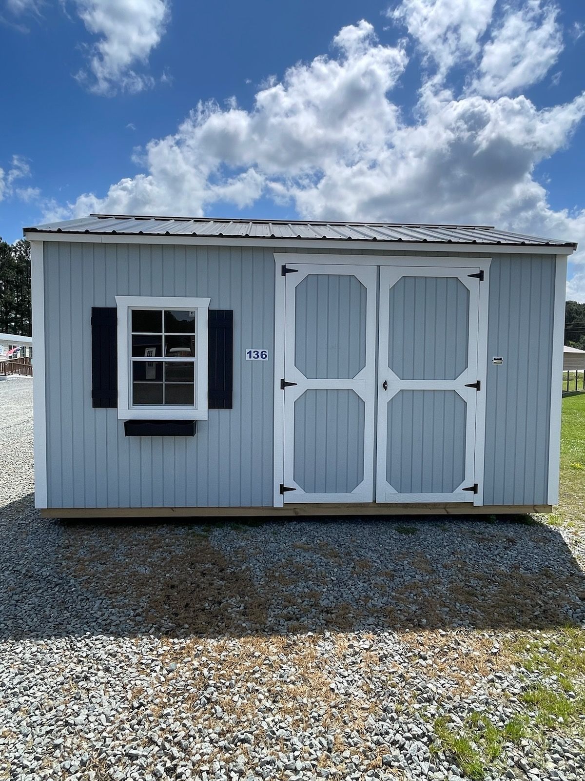 Light blue shed with white-trimmed doors, window, and dark shutters, set on gravel, under a cloudy sky.