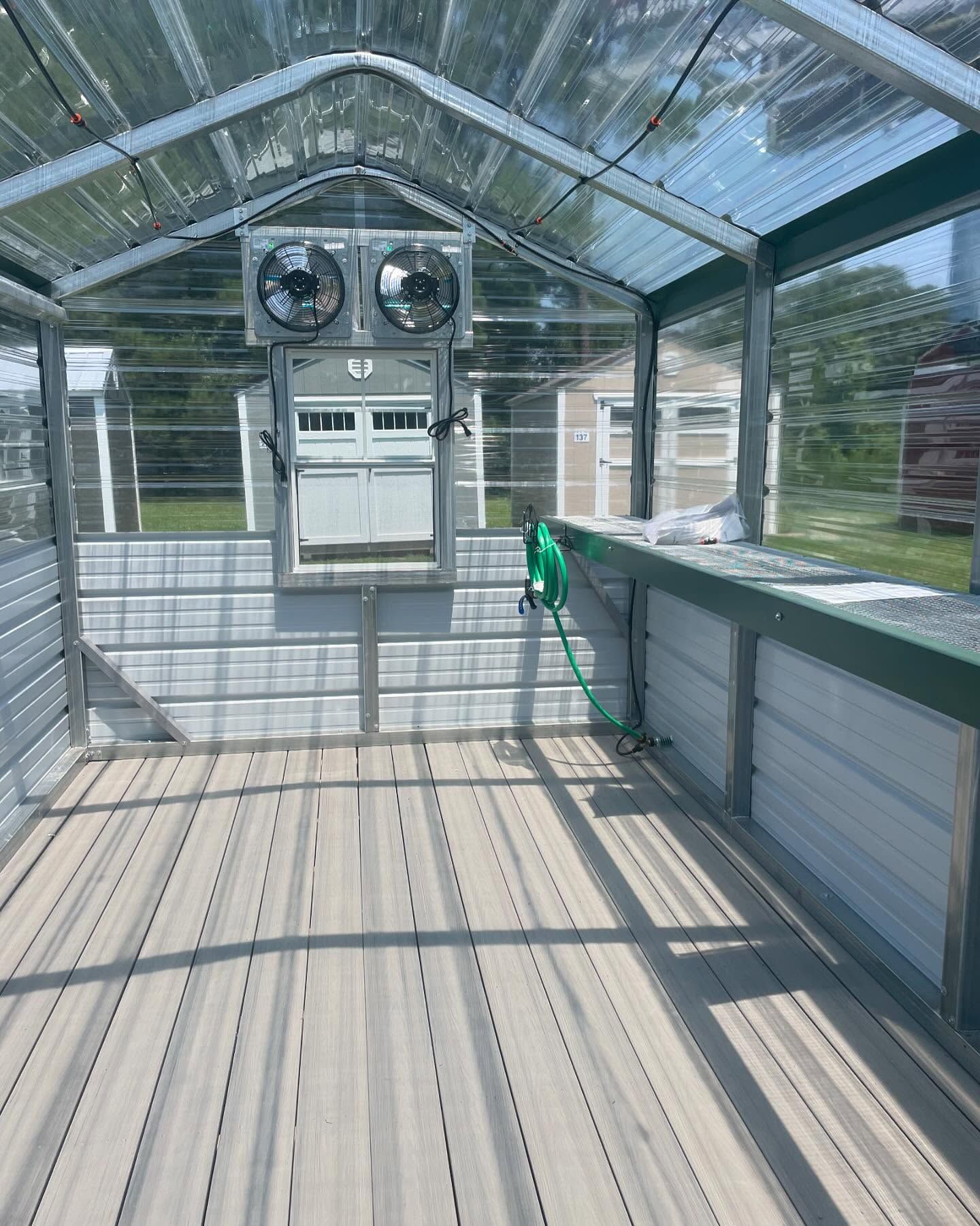 Interior of a greenhouse with a wooden floor, white and green walls, and two fans mounted on the back wall.